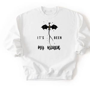 Puede incluir: Sudadera blanca con un dise&ntilde;o de drag&oacute;n negro y el texto "IT'S BEEN MY HONOR". El drag&oacute;n tiene las alas extendidas y est&aacute; posicionado encima del texto. La sudadera tiene mangas largas y cuello redondo.