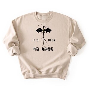 Puede incluir: Sudadera beige con el texto "IT'S BEEN my honor" y un gr&aacute;fico de drag&oacute;n negro. El drag&oacute;n tiene las alas extendidas y est&aacute; colocado encima del texto. La sudadera tiene mangas largas y cuello redondo.