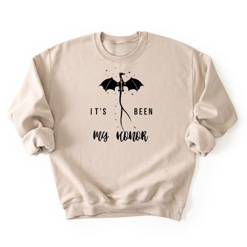 Puede incluir: Sudadera beige con el texto "IT'S BEEN my honor" y un gr&aacute;fico de drag&oacute;n negro. El drag&oacute;n tiene las alas extendidas y est&aacute; colocado encima del texto. La sudadera tiene mangas largas y cuello redondo.