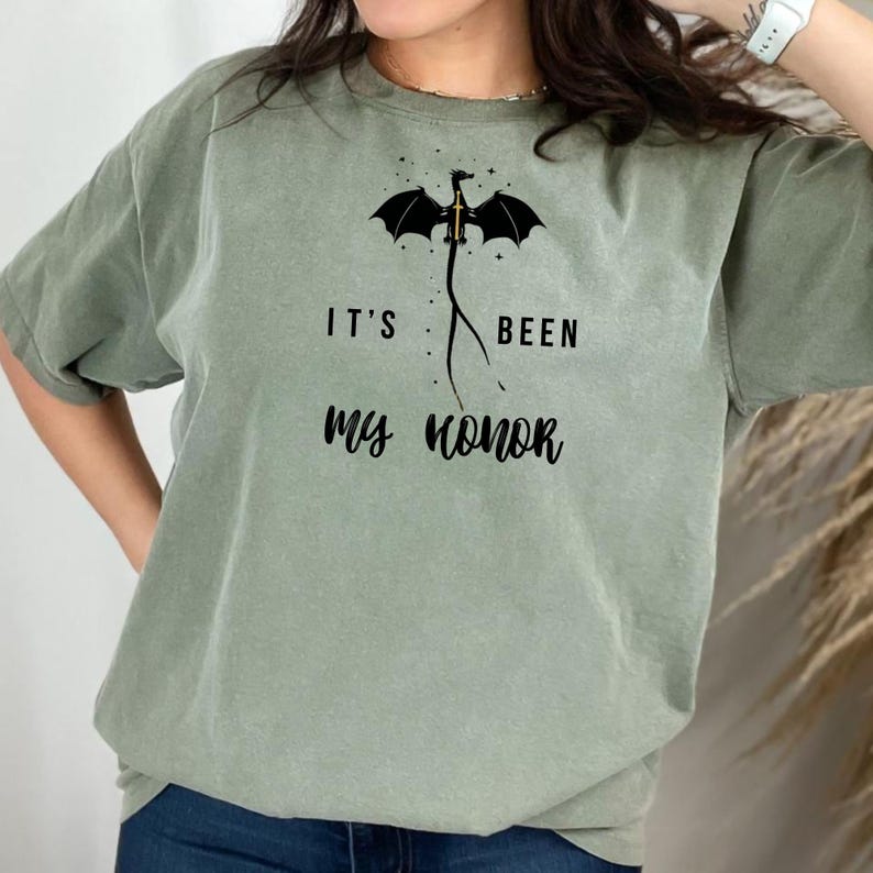 Puede incluir: Camiseta verde salvia con un gr&aacute;fico de drag&oacute;n negro y el texto "IT'S BEEN MY HONOR". El drag&oacute;n tiene alas extendidas y una espada. La camiseta es de corte holgado.