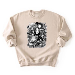 May include: A beige sweatshirt featuring a black and white graphic of various animated characters. The design includes figures like Totoro and No-Face, creating a collage-like composition. The sweatshirt has long sleeves and a crew neck.