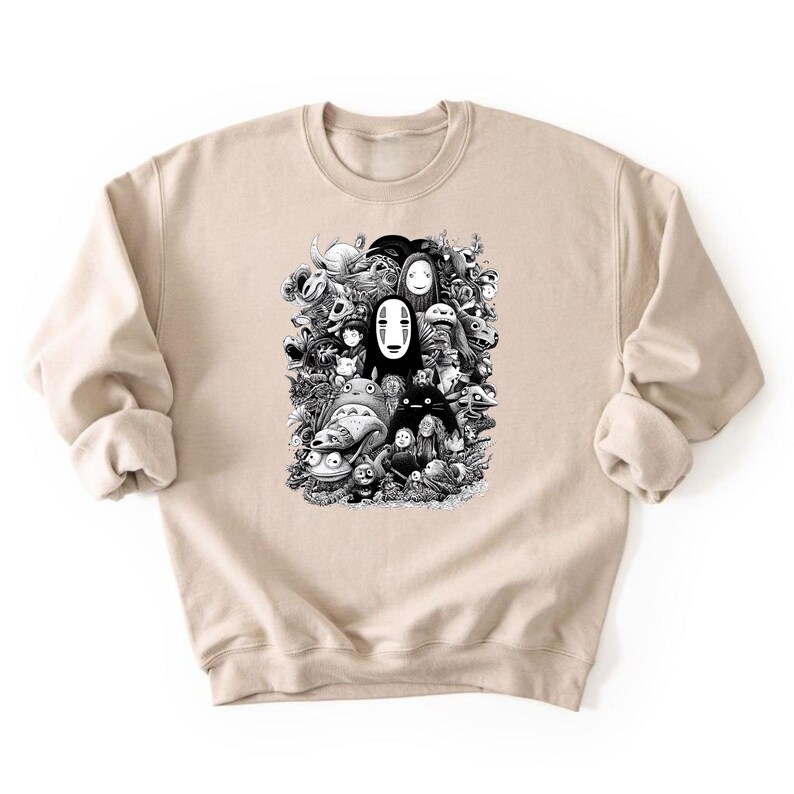 May include: A beige sweatshirt featuring a black and white graphic of various animated characters. The design includes figures like Totoro and No-Face, creating a collage-like composition. The sweatshirt has long sleeves and a crew neck.