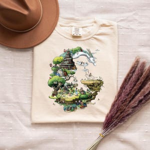 May include: Cream-colored t-shirt featuring a colorful illustration of a fantasy landscape with a dragon, buildings, and characters. A brown hat and dried flowers are in the background. The shirt is made of a soft material.