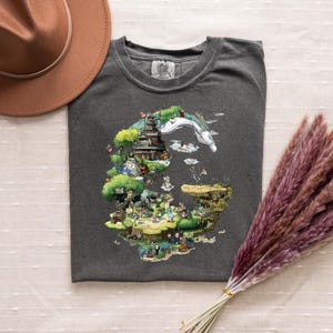 May include: Dark gray t-shirt featuring a colorful illustration of a fantasy landscape with a dragon, buildings, and characters. The shirt has a crew neck and short sleeves. A brown hat and dried flowers are in the background.