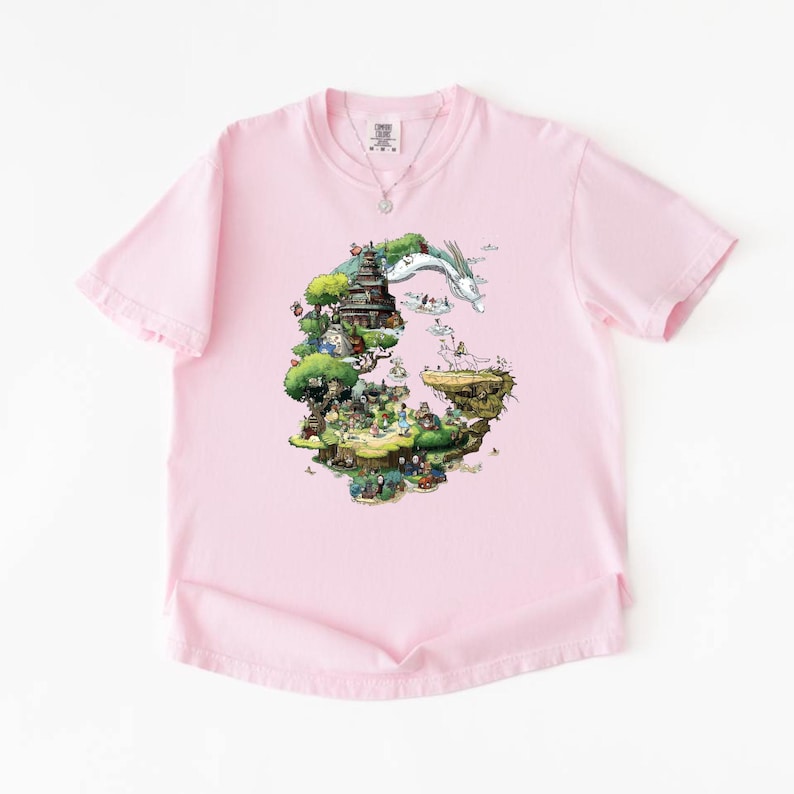 May include: A light pink t-shirt featuring a colorful graphic print. The print depicts a detailed fantasy landscape with a pagoda, trees, and figures, with a white dragon flying above. A silver necklace is visible at the neckline.
