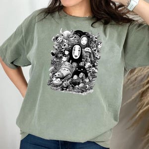 May include: Sage green t-shirt featuring a black and white graphic of various animated characters. The design includes a central figure with a white mask and other whimsical creatures. The shirt is made of a soft material.