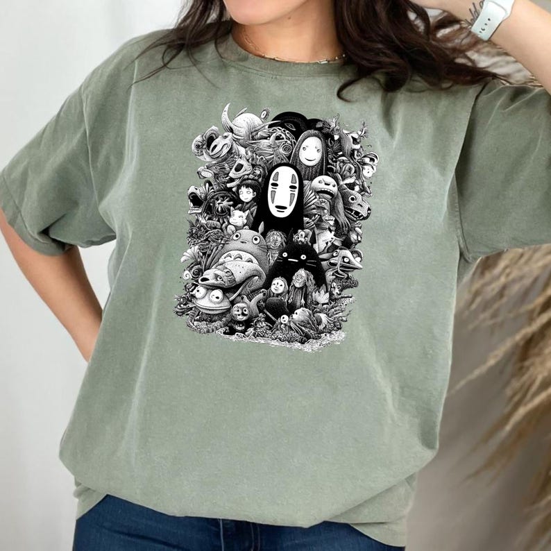 May include: Sage green t-shirt featuring a black and white graphic of various animated characters. The design includes a central figure with a white mask and other whimsical creatures. The shirt is made of a soft material.