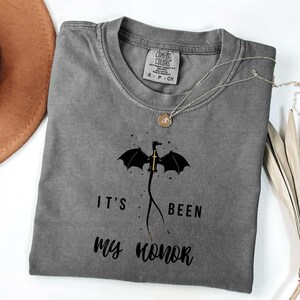 Puede incluir: Una camiseta gris Comfort Colors con un dise&ntilde;o de drag&oacute;n negro y una espada dorada, con el texto "IT'S BEEN MY HONOR". Un collar plateado con un colgante redondo est&aacute; sobre la camiseta.