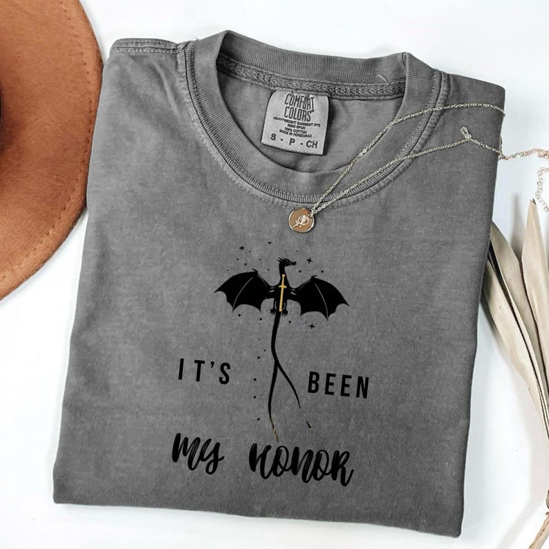 Puede incluir: Una camiseta gris Comfort Colors con un dise&ntilde;o de drag&oacute;n negro y una espada dorada, con el texto "IT'S BEEN MY HONOR". Un collar plateado con un colgante redondo est&aacute; sobre la camiseta.