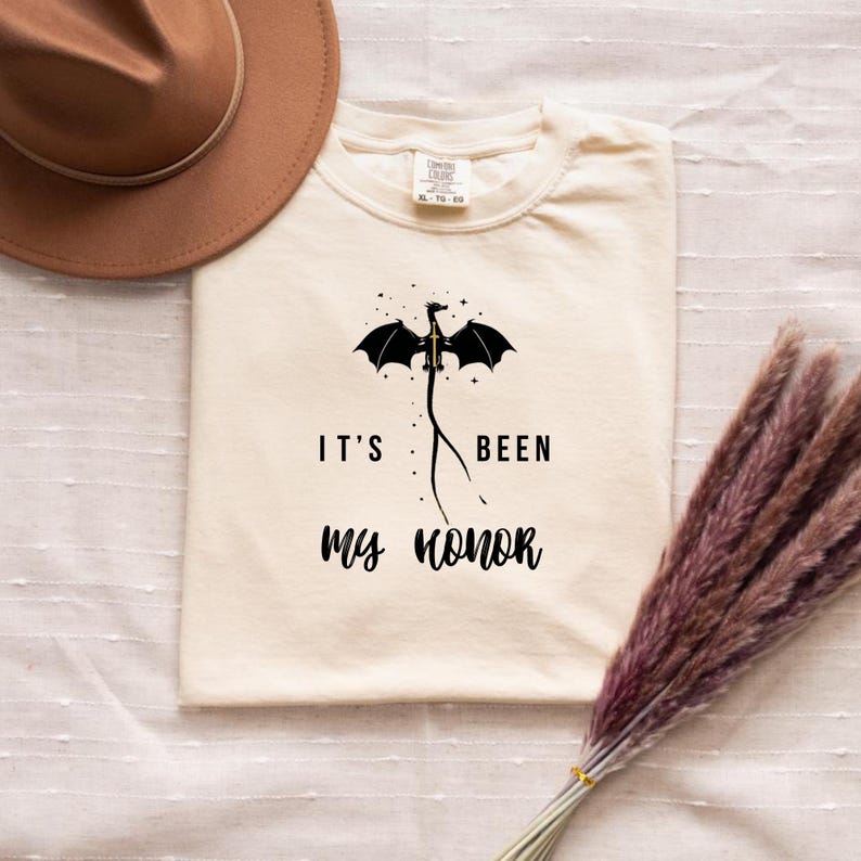 Puede incluir: Camiseta color crema con el texto "IT'S BEEN MY HONOR" y un dise&ntilde;o de drag&oacute;n negro. Un sombrero marr&oacute;n y un ramo de flores secas est&aacute;n junto a la camiseta.