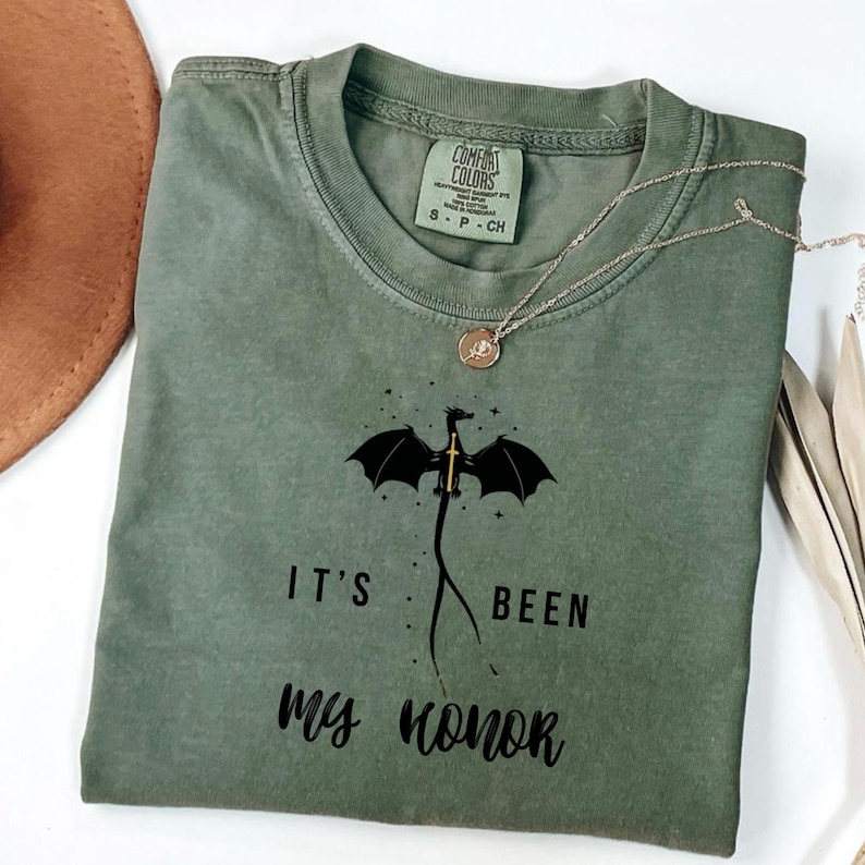 Puede incluir: Camiseta verde oliva con un gr&aacute;fico de drag&oacute;n y el texto "IT'S BEEN my honor". La camiseta tiene cuello redondo y mangas cortas. Un collar plateado con un colgante est&aacute; drapeado sobre la camiseta.