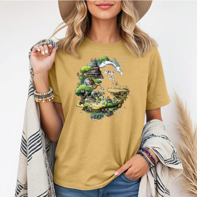 May include: Mustard yellow t-shirt featuring a colorful graphic of a fantasy landscape with a white dragon and various characters. The design includes trees, buildings, and figures, creating a whimsical scene. The shirt is made of a soft material.