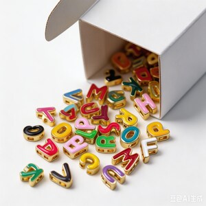 May include: A collection of small, gold-toned metal alphabet beads with colorful enamel centers in shades of red, pink, yellow, green, blue, purple, black, and white, spilling out from an open white cardboard box onto a plain white surface.
