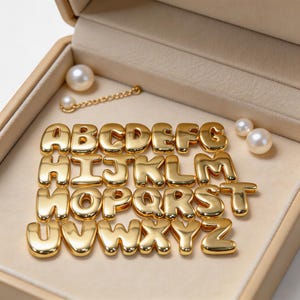 May include: A collection of twenty-six gold-toned metal alphabet letter charms, each featuring a rounded, puffy design. The set includes letters A through Z, accompanied by three white spherical pearls and a short gold-toned metal chain link.
