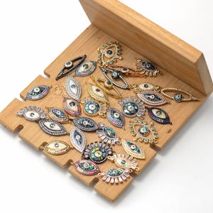 May include: A collection of eye-shaped jewelry charms in various metal finishes including gold, silver, and black. Each charm features iridescent abalone shell centers, clear crystal accents, and colorful pavé stones in a variety of eye designs.