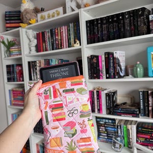 May include: A pink quilted book sleeve with a pattern of books, coffee cups, and glasses. The sleeve has an orange trim and the text "Just 1 More Chapter" and "Read More Books". A tablet with the text "Sarah J. Maas Throne" is inside the sleeve.