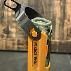 May include: A portable speaker with a camouflage pattern, secured in a custom orange and black holder. The holder has a handle and the text "TRAIL EDITION" printed on the side.