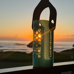May include: A teal cylindrical lantern with a honeycomb pattern, suspended by a black handle. The text "SEAGLASS EDITION" is printed vertically on the side. The setting sun shines through the lantern's openings, with a view of the ocean in the background.