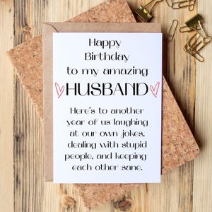 May include: A white rectangular greeting card with black text that reads: Happy Birthday to my amazing HUSBAND. The text is flanked by two small pink hearts. The card features a humorous message about laughing at jokes and keeping each other sane.