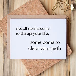 May include: A white rectangular greeting card with black text that reads: not all storms come to disrupt your life, some come to clear your path. The card is placed inside a light brown kraft paper envelope, resting on a cork square.