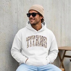 May include: A white hooded sweatshirt featuring the word TITANS in an arched, camouflage-patterned font across the chest. The hoodie includes a front kangaroo pocket, long sleeves, and ribbed cuffs, providing a casual and functional garment.