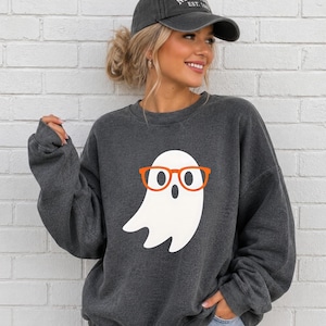 Può includere: Una felpa girocollo grigio antracite scuro con una grande grafica di un fantasma bianco che indossa occhiali arancioni vivaci. La felpa è abbinata a un berretto da baseball grigio scuro con la scritta NEW YORK EST. 1624 ricamata in bianco.