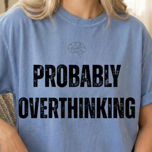 May include: A light blue t-shirt with the words "PROBABLY OVERTHINKING" in large, black, distressed font. Above the text is a small, black line drawing of a brain. The shirt has rolled-up sleeves.