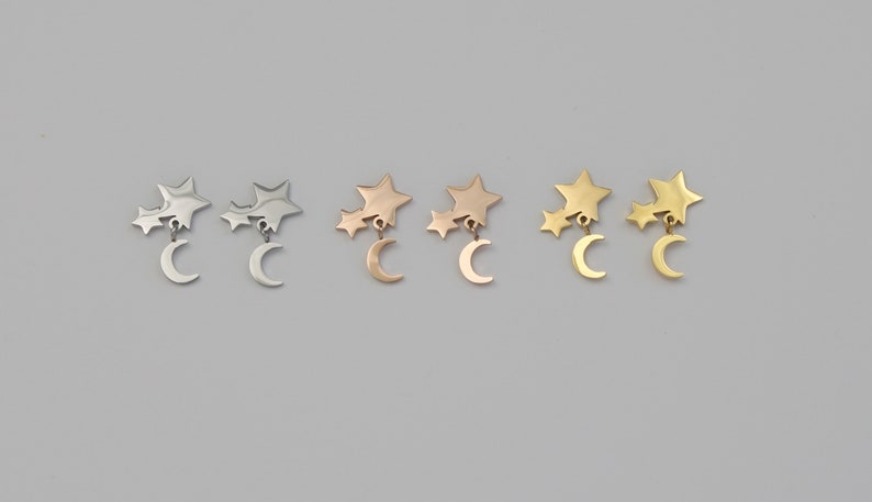 May include: Three pairs of earrings featuring a crescent moon charm with two star charms dangling from it. The earrings are shown in silver, rose gold, and gold.