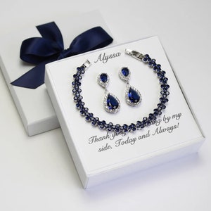 May include: A jewelry set featuring a bracelet and matching earrings with dark blue gemstones and silver-colored metal. The set is displayed on a white box with the text "Alyssa" and "Thank you for standing by my side, Today and Always!" A navy blue ribbon adorns the box.