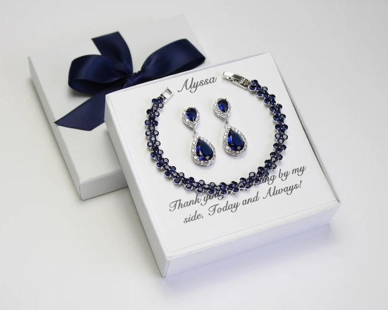 May include: A jewelry set featuring a bracelet and matching earrings with dark blue gemstones and silver-colored metal. The set is displayed on a white box with the text "Alyssa" and "Thank you for standing by my side, Today and Always!" A navy blue ribbon adorns the box.