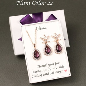 May include: A white gift box with a purple ribbon and a clear lid. Inside the box is a rose gold necklace and earrings set with a teardrop-shaped purple gemstone and clear rhinestones. The box has the text "Olivia" and "Thank you for standing by my side, Today and Always!" printed on it.