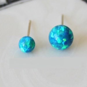 Puede incluir: Un par de pendientes de bot&oacute;n de &oacute;palo con un acabado iridiscente azul y verde. Los pendientes est&aacute;n sobre una superficie blanca.