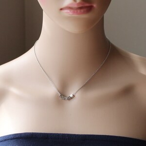 Puede incluir: Un collar de cadena de plata con un peque&ntilde;o colgante hecho de varios cubos de plata. El colgante tiene la forma de una palabra.