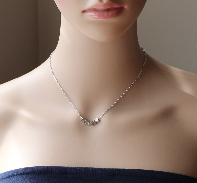 Puede incluir: Un collar de cadena de plata con un peque&ntilde;o colgante hecho de varios cubos de plata. El colgante tiene la forma de una palabra.