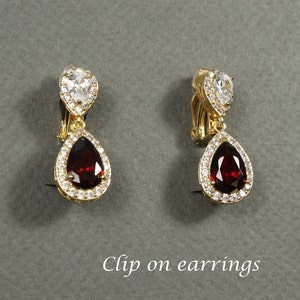 May include: A pair of gold-toned clip-on earrings. Each earring features a pear-shaped, deep red gemstone surrounded by small, clear crystals. A smaller, heart-shaped clear crystal is at the top. The text "Clip on earrings" is at the bottom.