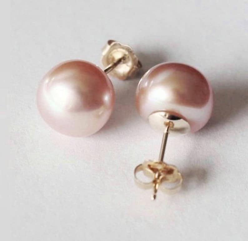 May include: A pair of stud earrings featuring round, light pink pearls with a smooth, lustrous finish. Each pearl is mounted on a gold-toned metal post with a matching gold-toned butterfly clutch backing, showing the hardware clearly.