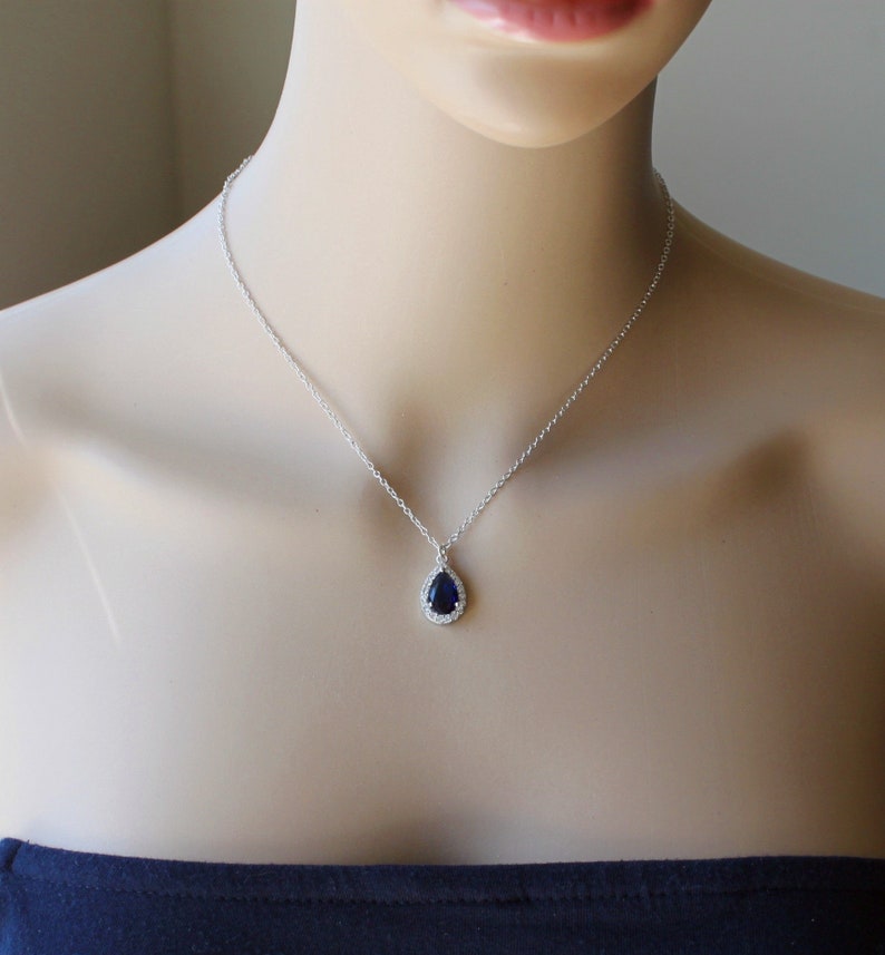 May include: A silver necklace with a teardrop-shaped pendant featuring a dark blue gemstone surrounded by smaller clear gemstones.
