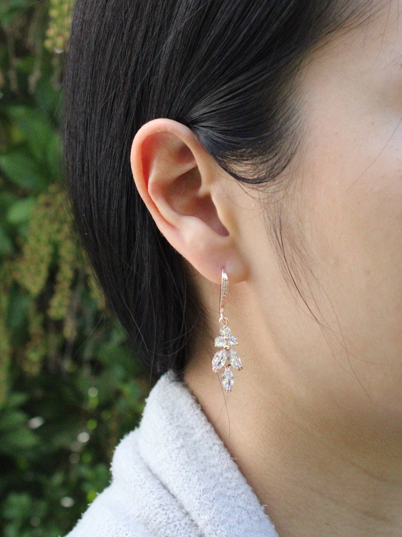 May include: Rose gold dangle earrings with clear cubic zirconia stones. The earrings feature a cluster of stones in a leaf-like design.
