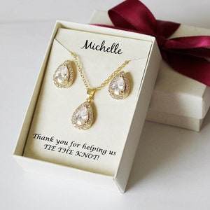 Puede incluir: Una caja de regalo blanca con una cinta roja, que contiene un conjunto de collar y pendientes de oro con piedras transparentes en forma de pera. La caja tiene un mensaje personalizado que dice "Michelle" y "&iexcl;Gracias por ayudarnos a ATAR EL NUDO!"