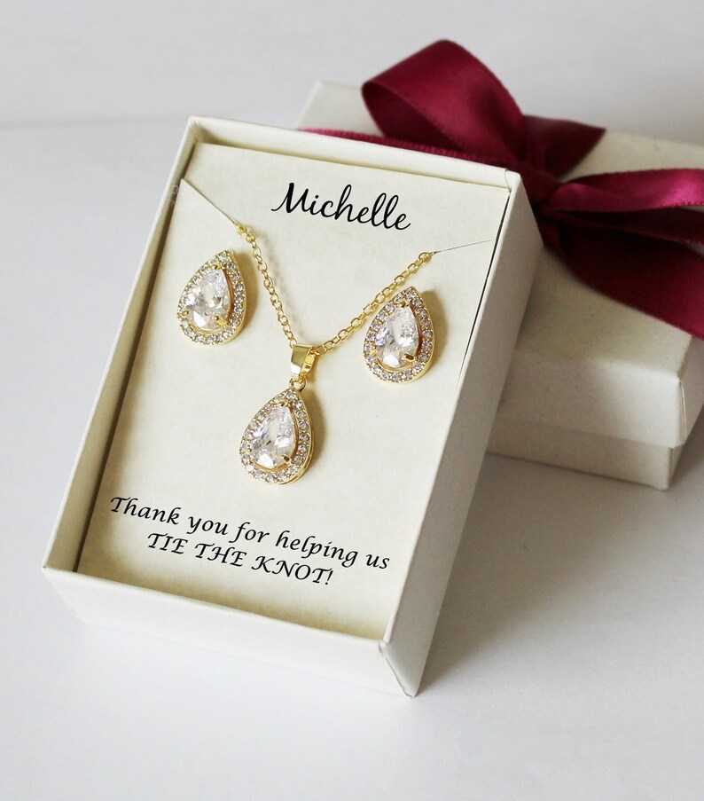 Puede incluir: Una caja de regalo blanca con una cinta roja, que contiene un conjunto de collar y pendientes de oro con piedras transparentes en forma de pera. La caja tiene un mensaje personalizado que dice "Michelle" y "&iexcl;Gracias por ayudarnos a ATAR EL NUDO!"