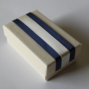 May include: A small, rectangular box with a cream-colored exterior and a navy blue and white ribbon design.