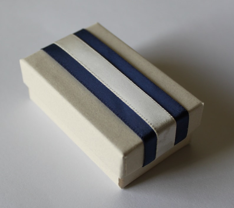 May include: A small, rectangular box with a cream-colored exterior and a navy blue and white ribbon design.