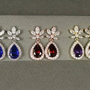 May include: Three pairs of dangle earrings with a teardrop-shaped stone surrounded by smaller stones. The earrings are available in silver, rose gold, and gold with blue, red, and purple stones.