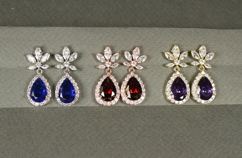 May include: Three pairs of dangle earrings with a teardrop-shaped stone surrounded by smaller stones. The earrings are available in silver, rose gold, and gold with blue, red, and purple stones.