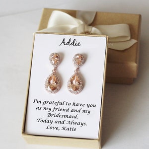 May include: A small gift box with a white card inside. The card has the name "Addie" written on it and a message that says "I'm grateful to have you as my friend and my Bridesmaid. Today and Always. Love, Katie". The box contains a pair of rose gold earrings with teardrop-shaped crystals.