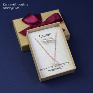May include: A gift box with a burgundy ribbon and a personalized message for Lauren. Inside the box is a rose gold necklace and earrings set with pearl accents. The message reads "Thank you for being my Bridesmaid!"