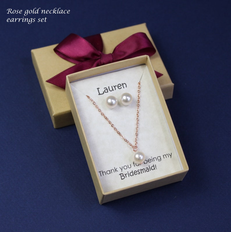 May include: A gift box with a burgundy ribbon and a personalized message for Lauren. Inside the box is a rose gold necklace and earrings set with pearl accents. The message reads "Thank you for being my Bridesmaid!"