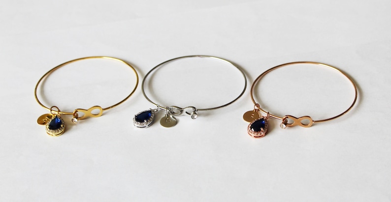 May include: Three delicate bangle bracelets with infinity charm and a teardrop-shaped pendant with a blue gemstone. The bracelets are available in gold, silver, and rose gold.