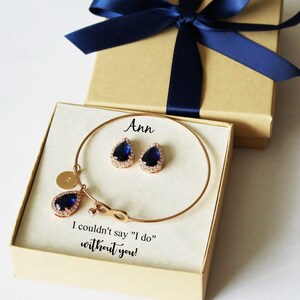May include: A gift box with a blue ribbon and a white card inside. The card says "Ann" and "I couldn't say "I do" without you!" The box contains a gold bracelet with a teardrop-shaped blue stone charm and matching earrings.