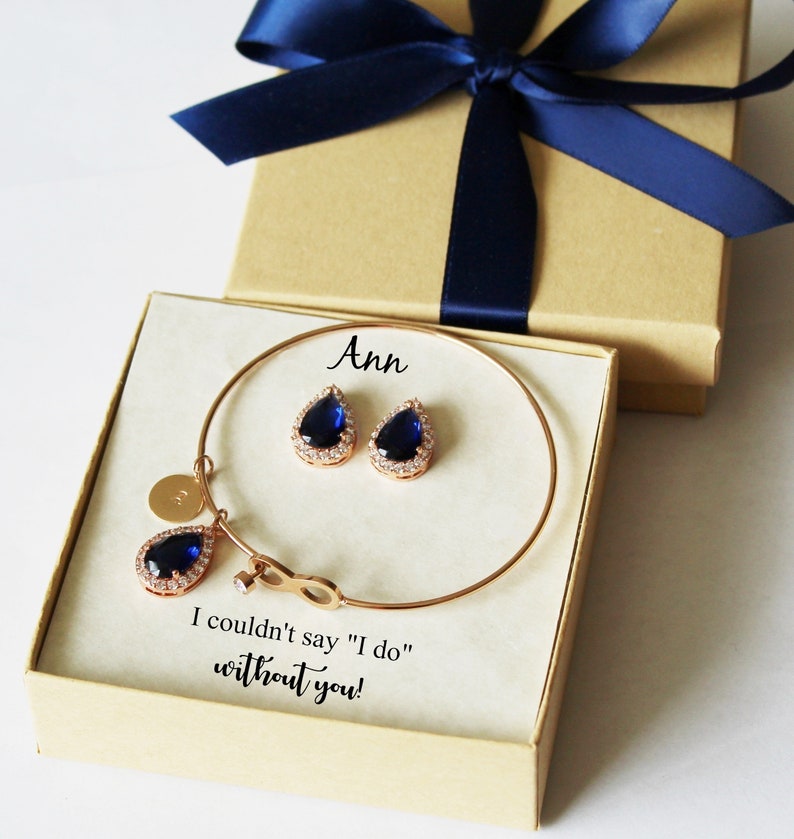 May include: A gift box with a blue ribbon and a white card inside. The card says "Ann" and "I couldn't say "I do" without you!" The box contains a gold bracelet with a teardrop-shaped blue stone charm and matching earrings.